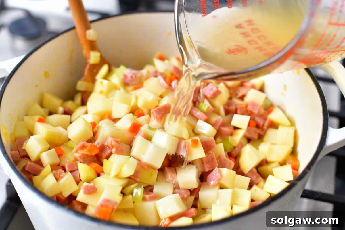 Chicken stock being poured into a pot filled with vegetables, potatoes, and ham for a soup recipe.