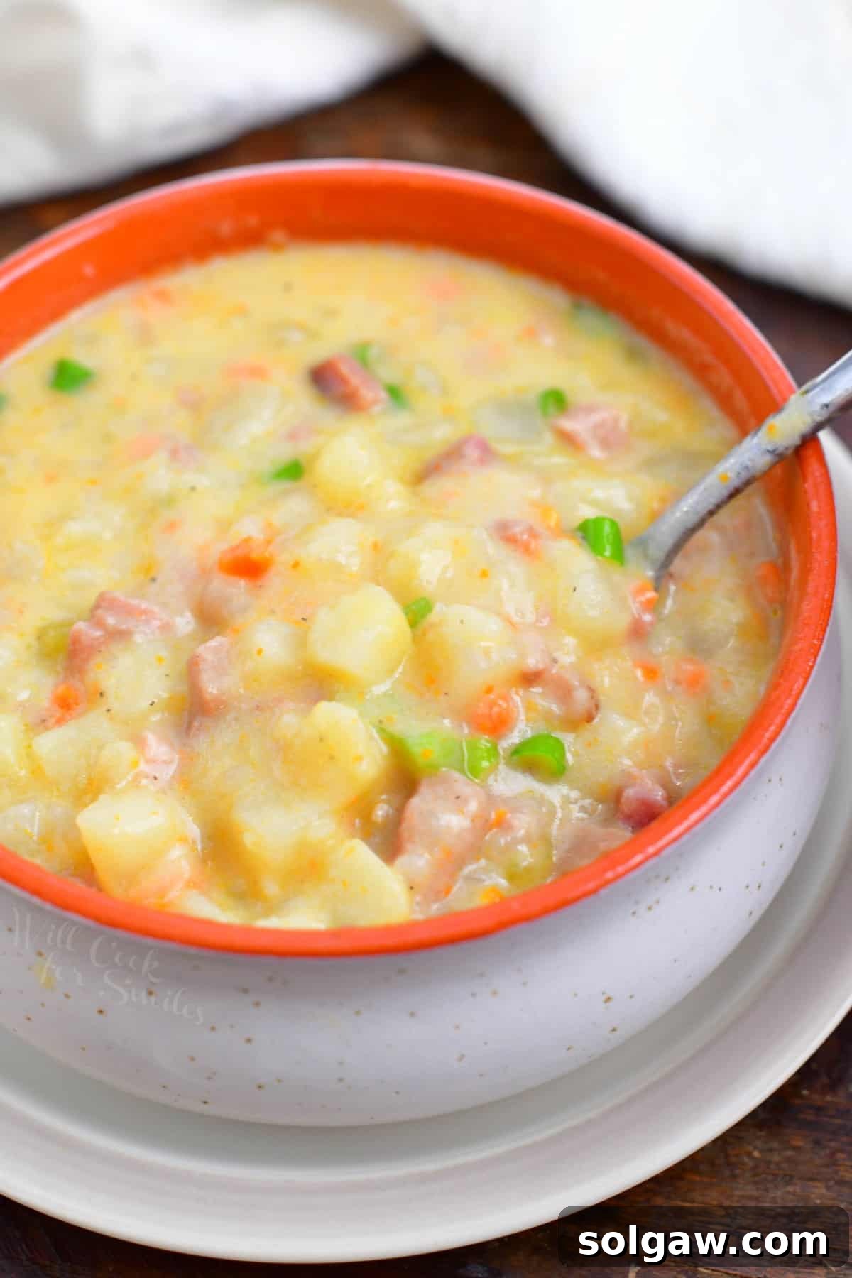 Overhead shot of a spoon in an orange bowl of creamy ham and potato soup, garnished and ready to eat.