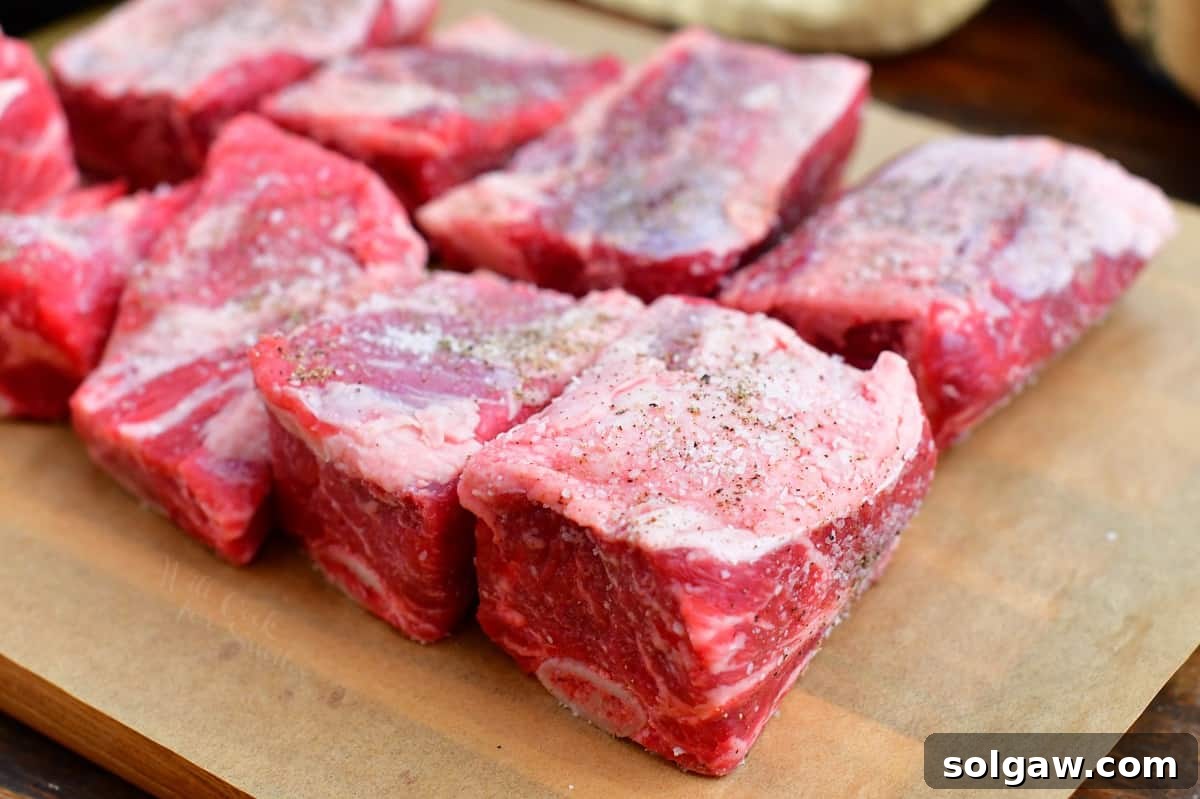 Uncooked beef short ribs seasoned with coarse salt and black pepper, resting on a wooden cutting board, ready for searing.