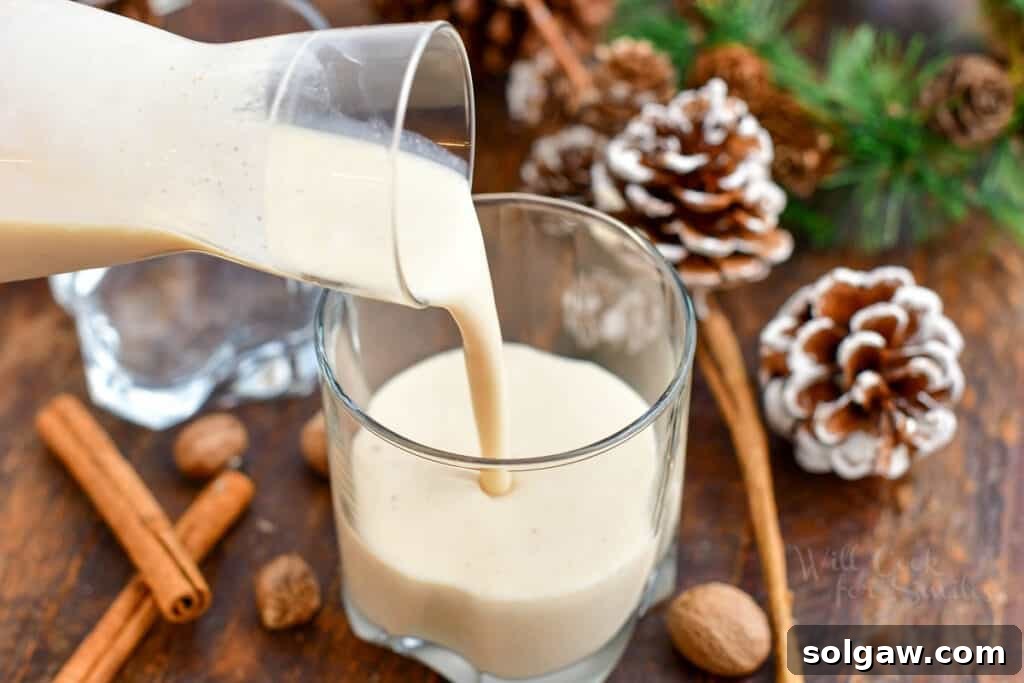 A close-up shot of a hand gracefully pouring freshly made Coquito from a clear glass pitcher into a classic Old Fashioned glass, ready for enjoyment.