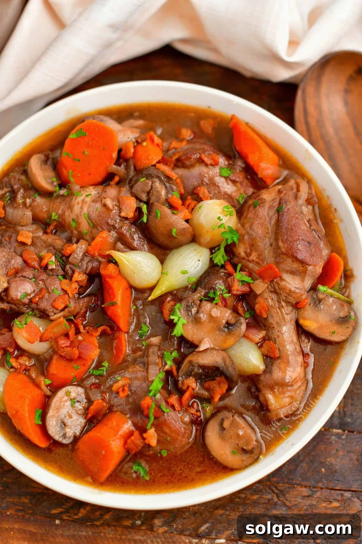 French Wine-Braised Chicken 9 Overhead view of a white bowl of Coq au Vin on a wooden surface, surrounded by a white cloth and wooden spoon, highlighting the finished presentation.