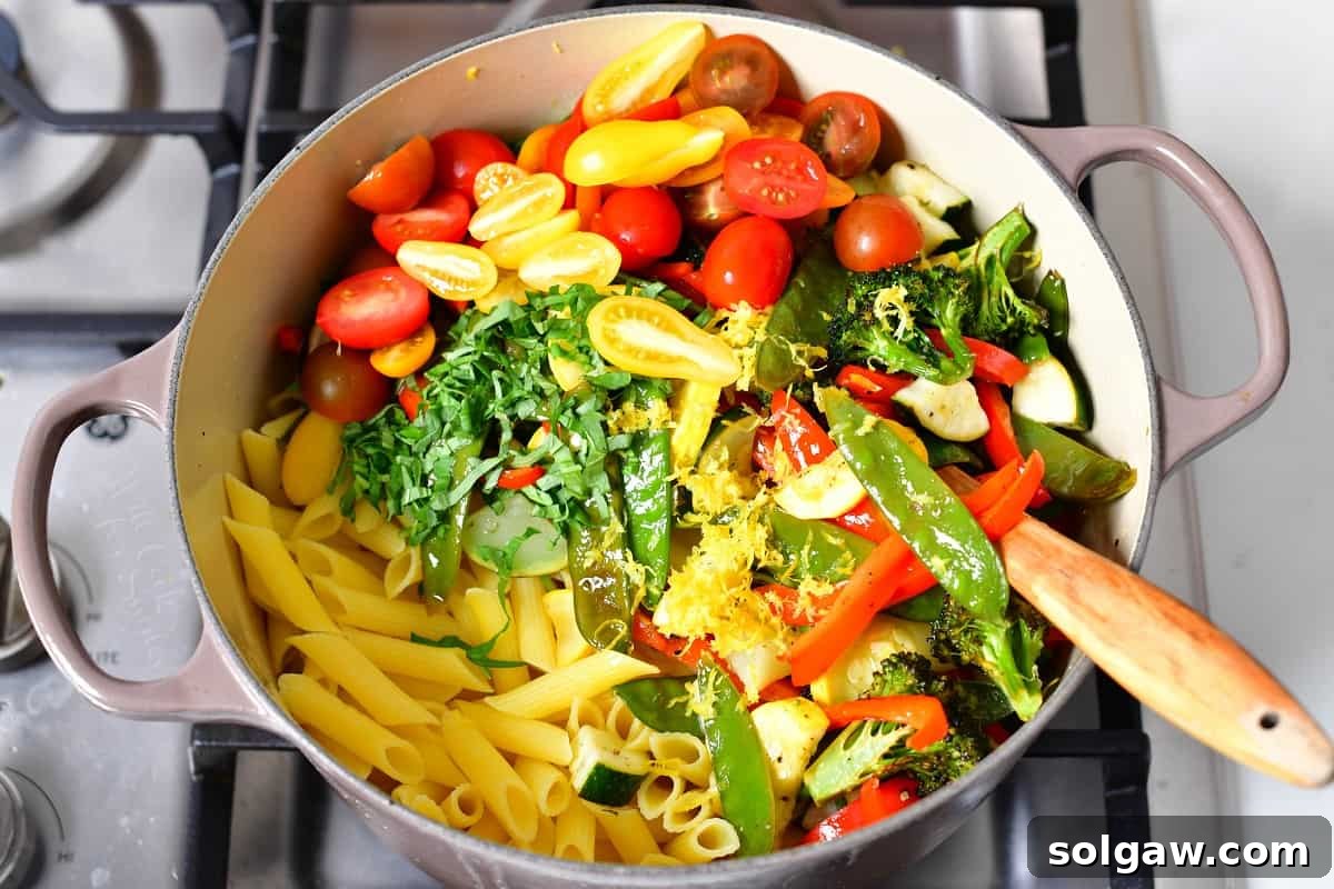 All the pasta primavera ingredients, including cooked pasta, roasted vegetables, and sauce, being tossed together in a Dutch oven.