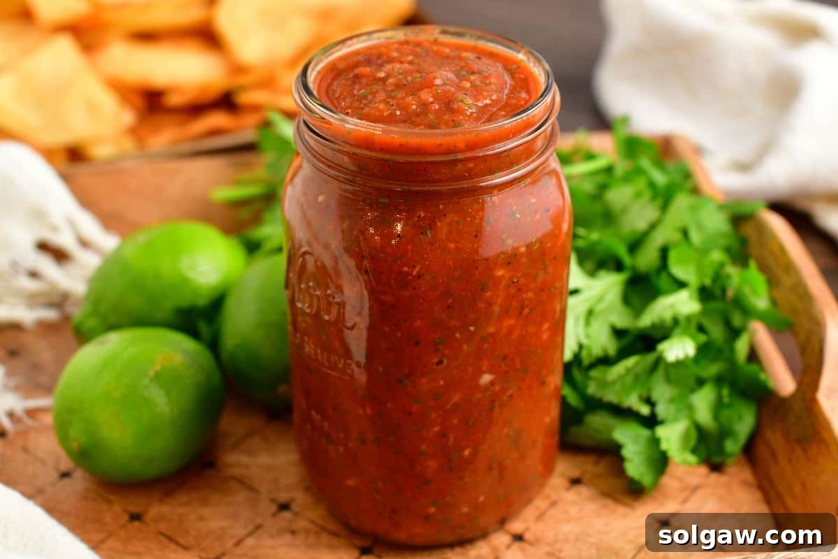 A glass jar filled with red salsa, surrounded by fresh limes and cilantro sprigs.