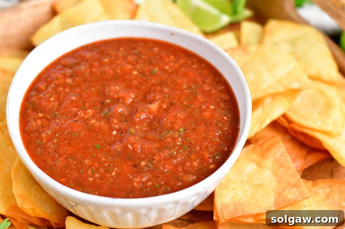 A bowl of vibrant red salsa is served next to a pile of golden tortilla chips on a white surface.