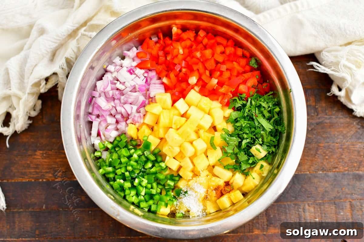 All the fresh ingredients for homemade pineapple salsa, including diced pineapple, red onion, bell pepper, and cilantro, are gathered in a large silver mixing bowl.