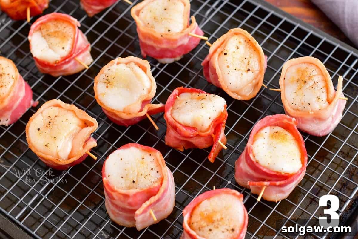 Scallops neatly wrapped in par-cooked bacon, secured with toothpicks, arranged on a baking rack before broiling.