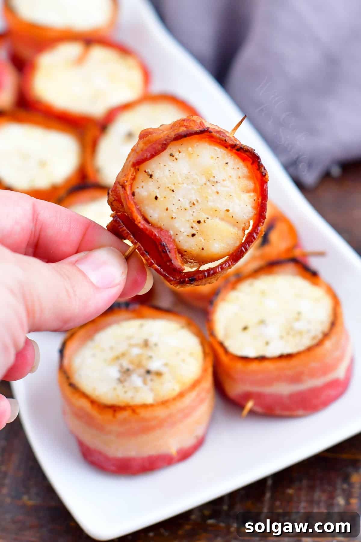 A single bacon wrapped scallop being lifted from a white plate, showcasing its crispy bacon and perfectly cooked interior.