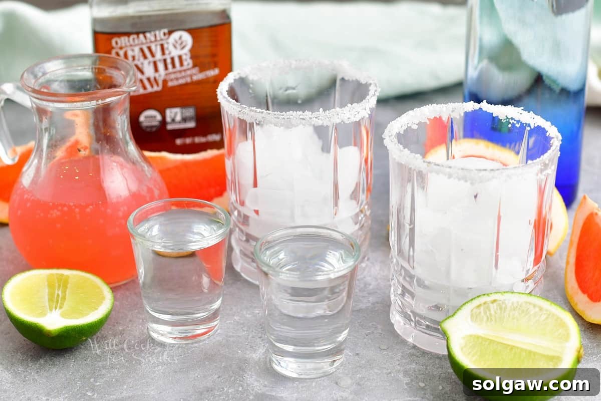 All the fresh ingredients for a Paloma cocktail laid out on a kitchen counter: limes, grapefruit soda, agave nectar, and tequila blanco.