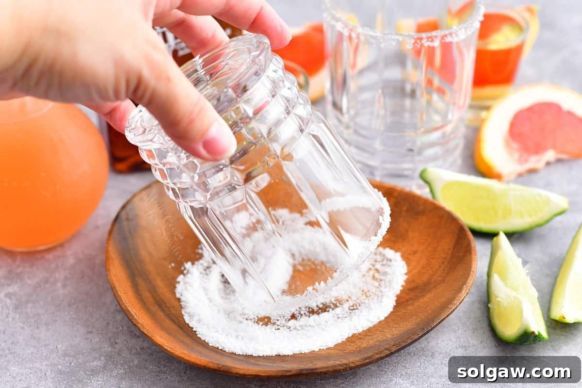 A hand is shown rimming a cocktail glass with coarse salt after moistening the edge with a lime wedge.