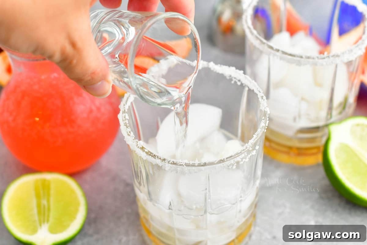 A shot of clear tequila blanco is being poured from a jigger into a salt-rimmed glass filled with ice.