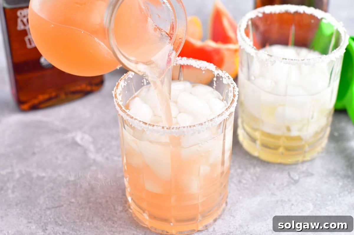 A clear glass of Paloma cocktail is being topped with grapefruit soda, showing the bubbles and vibrant pink color.