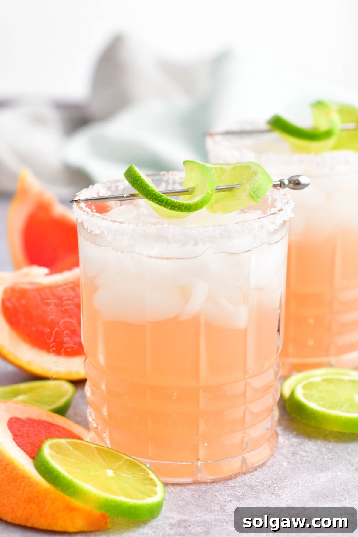 A vibrant pink Paloma cocktail, garnished with a fresh lime wedge, sits on a wooden surface.