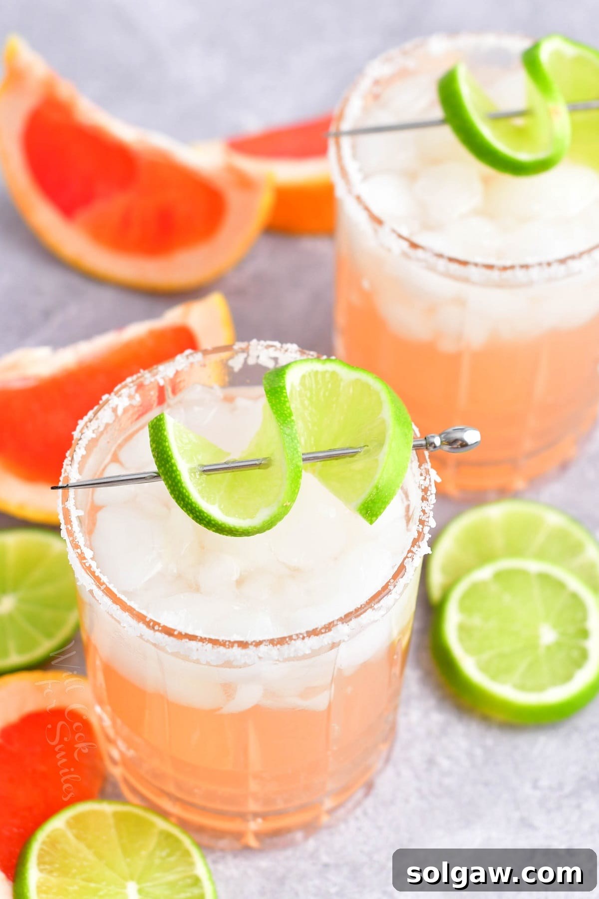 Two vibrant Paloma cocktails are shown, garnished with generous lime and grapefruit wedges, surrounded by more fresh citrus.