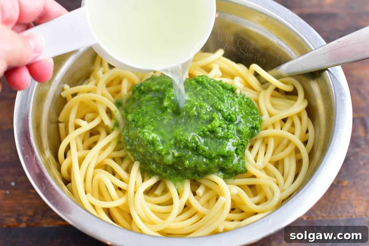 Freshly cooked pasta noodles in a spacious white bowl, generously topped with vibrant green homemade pesto sauce and a splash of reserved pasta cooking water, ready for mixing.