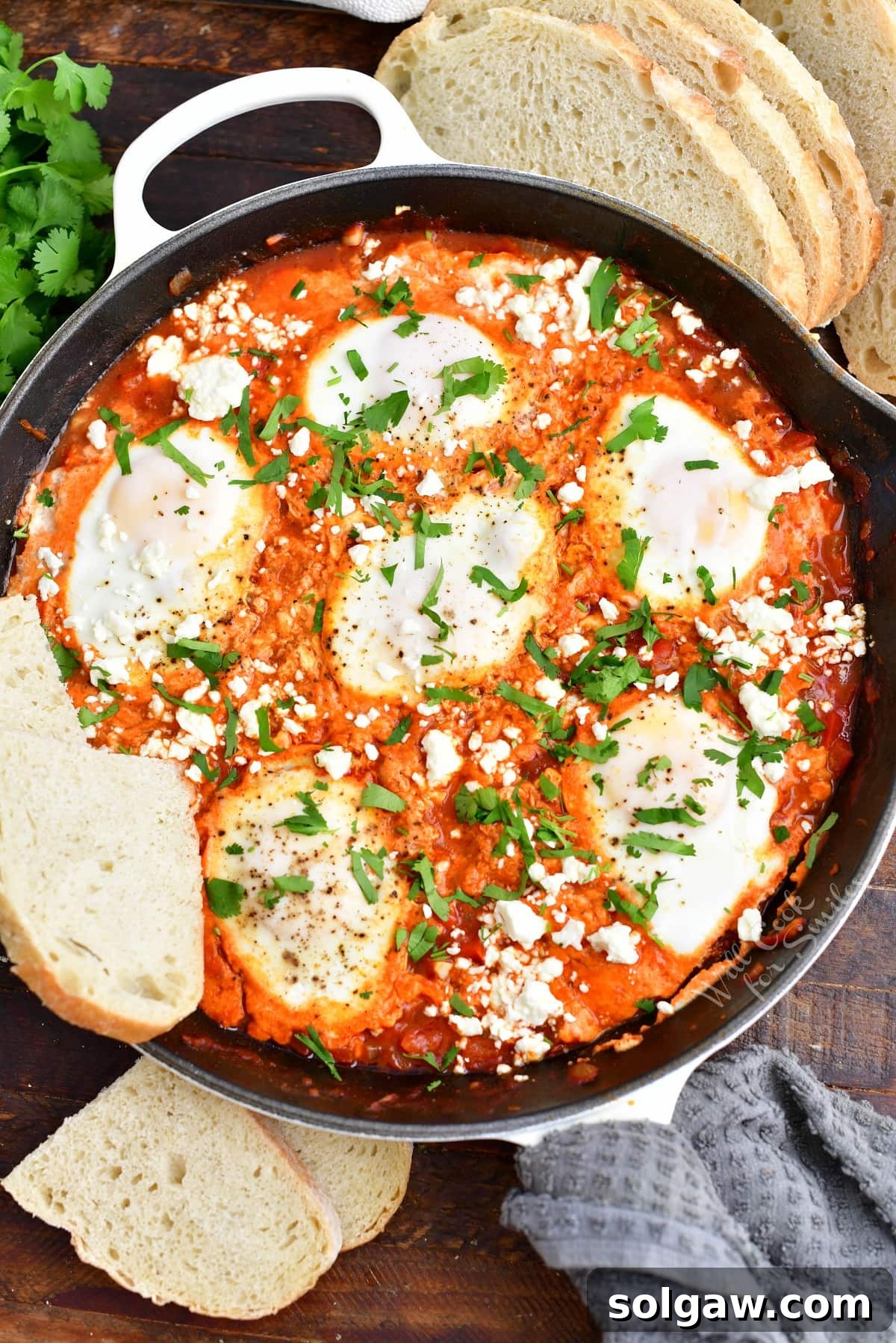 A few slices of rustic bread are placed next to a black skillet filled with vibrant red shakshuka, showcasing perfectly poached eggs and fresh herbs.