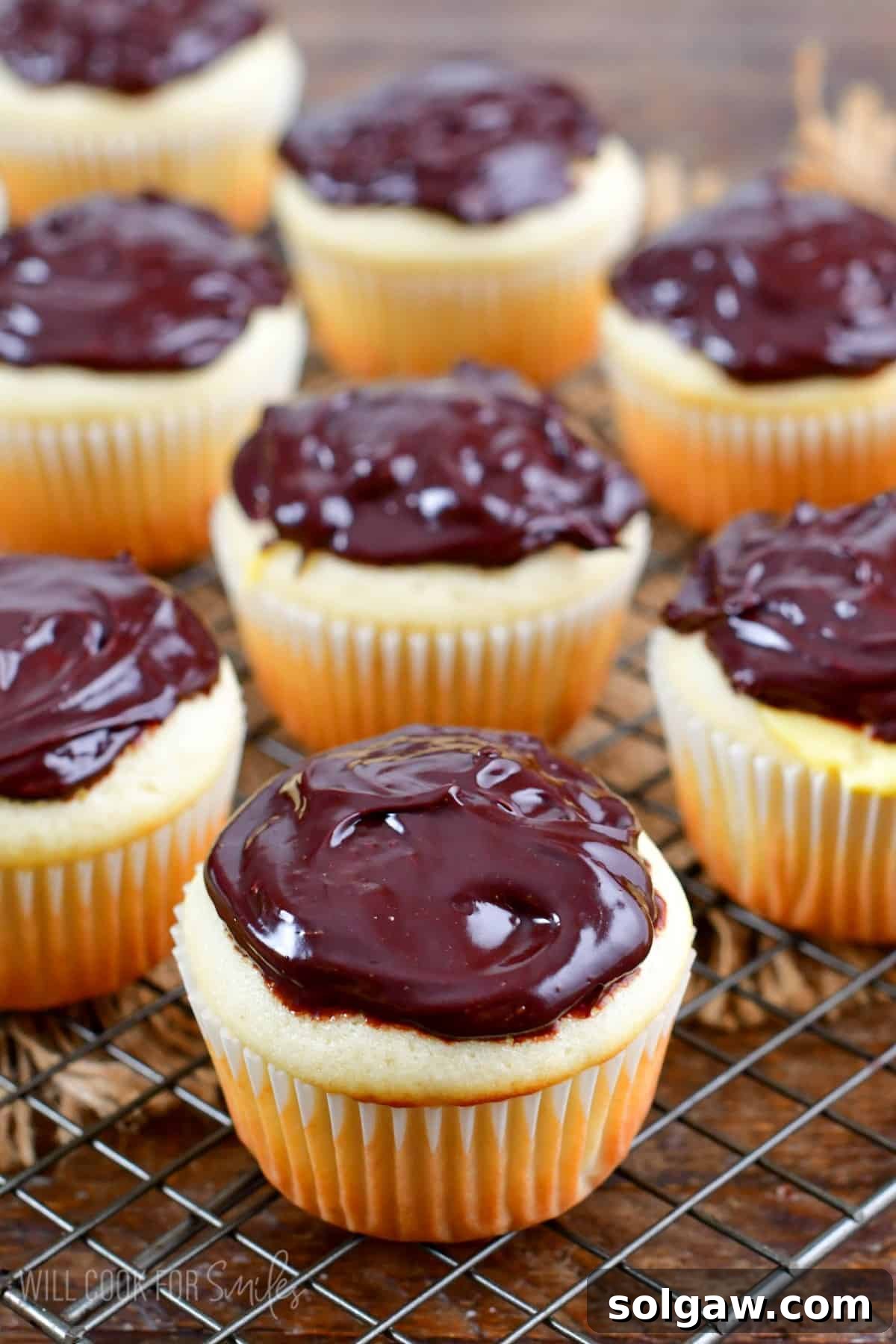 Boston Cream cupcakes on a wire rack with chocolate frosting on top on a wood surface.