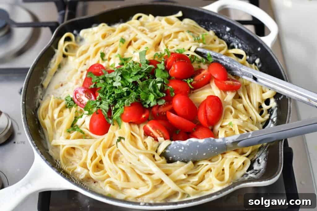 Fresh cherry tomatoes and green basil leaves are added to the creamy fettuccine pasta in a pan, ready to be tossed together.