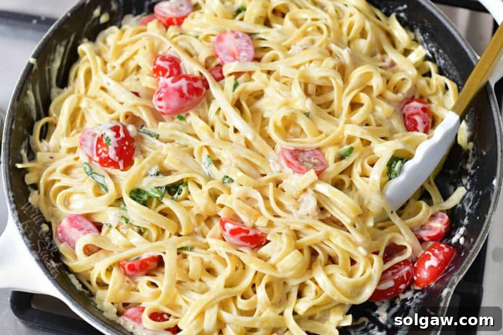 The finished dish: fettuccine pasta, cherry tomatoes, and basil thoroughly tossed in a rich Parmesan cream sauce, glistening in a cooking pan.