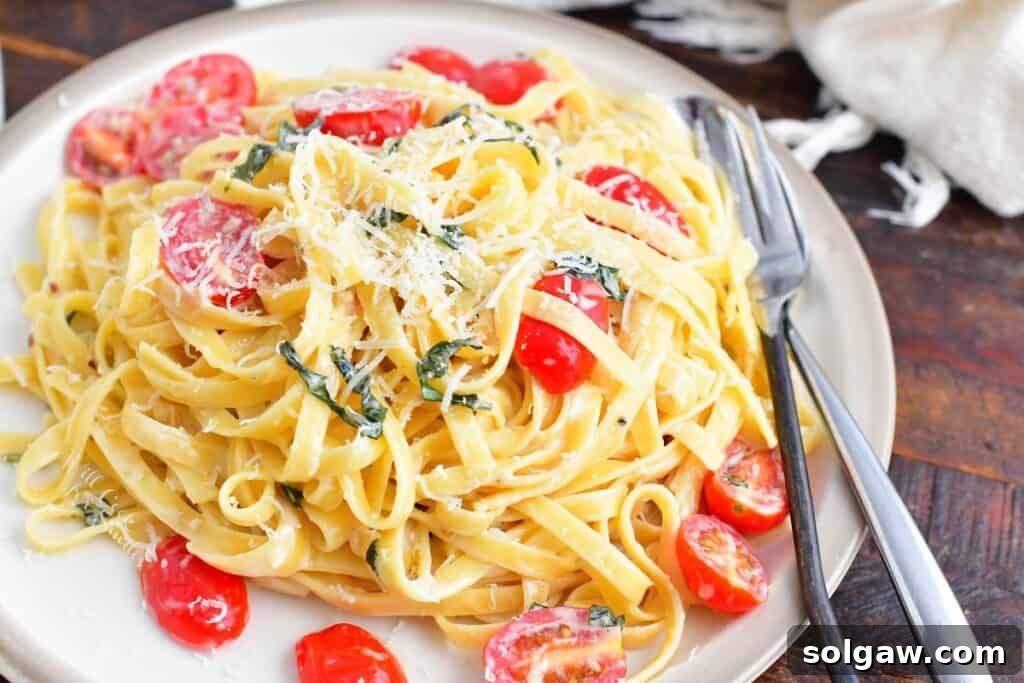 A beautifully plated serving of creamy fettuccine with cherry tomatoes and fresh basil, garnished with Parmesan, ready on a white plate with a fork and spoon.