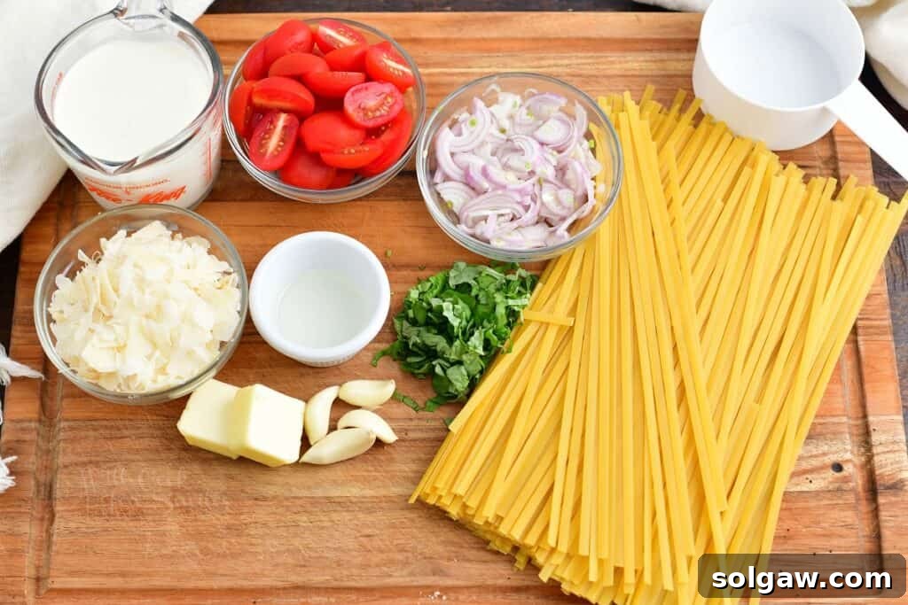 A cutting board displays fresh ingredients including pasta, cherry tomatoes, basil leaves, shallots, garlic cloves, butter, and olive oil, all ready for cooking creamy fettuccine.