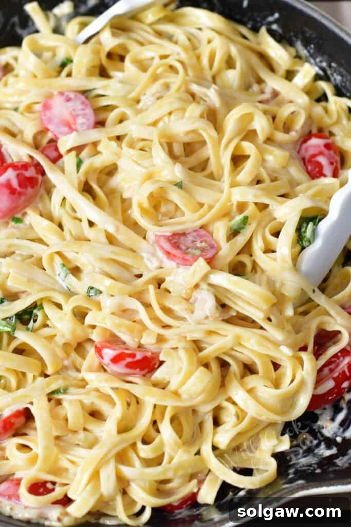 Closeup shot of pasta strands tossed in a rich cream sauce with halved cherry tomatoes and fresh basil leaves inside a large cooking pan.