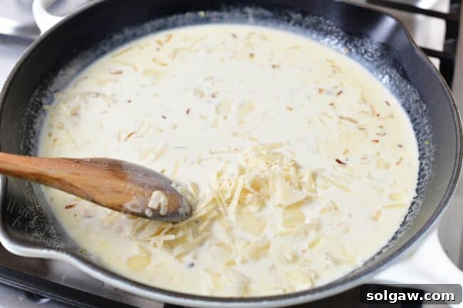 A close-up view of a spoon stirring shaved Parmesan cheese into a creamy white sauce in a pan, ensuring a smooth and even melt.