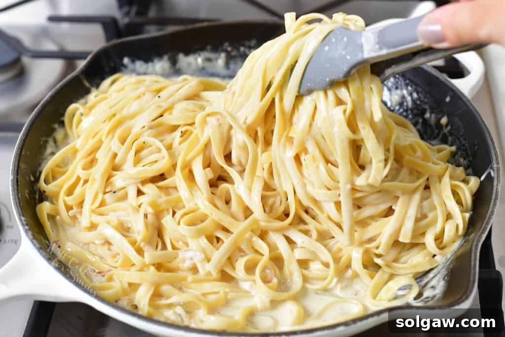 Tongs are used to expertly toss fettuccine pasta in a creamy white sauce within a large cooking pan, ensuring even coating.