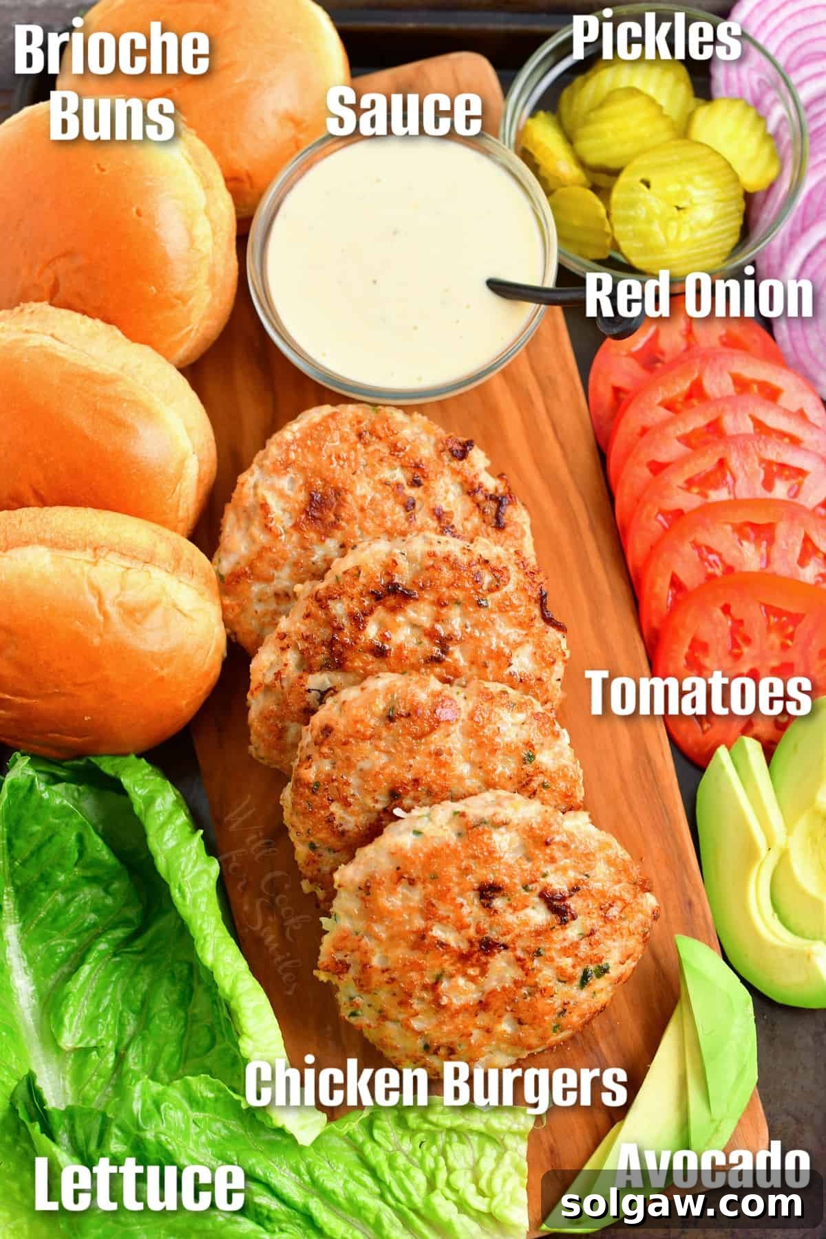Ultimate Chicken Burgers 8 Ingredients assembled to build a chicken burger, including a patty, brioche bun, lettuce, tomato, onion, pickles, and sauce on a cutting board.