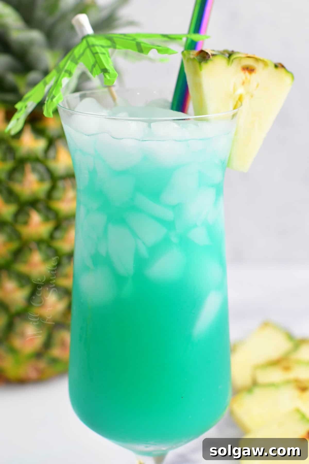 Close-up side view of a vibrant blue Hawaii cocktail in a hurricane glass, garnished with a fresh pineapple wedge and a small tropical umbrella, evoking a beach vacation feeling.