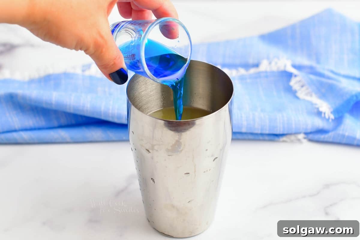 A hand pouring vibrant blue curacao into a cocktail shaker, beginning the process of mixing a Blue Hawaii cocktail.