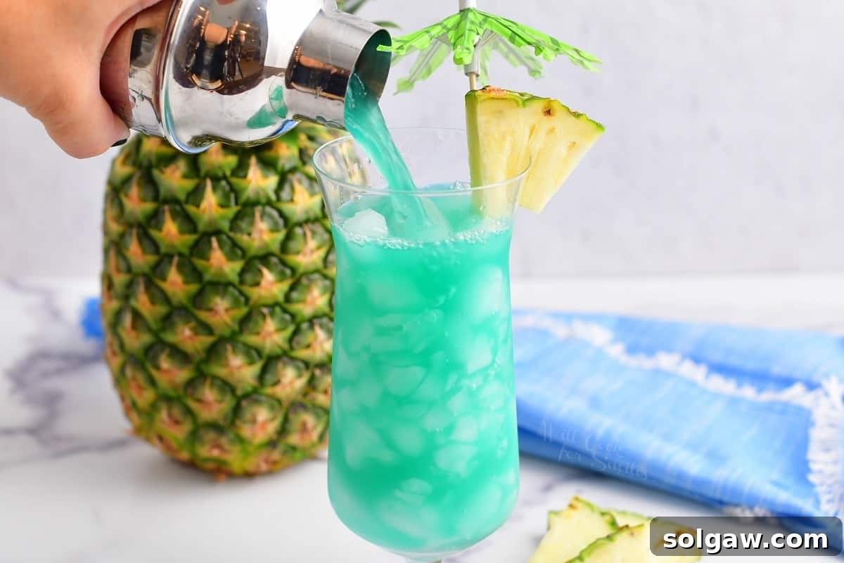 Vibrant blue Hawaii cocktail being poured from a shaker into a hurricane glass filled with crushed ice, showcasing the refreshing drink in action.
