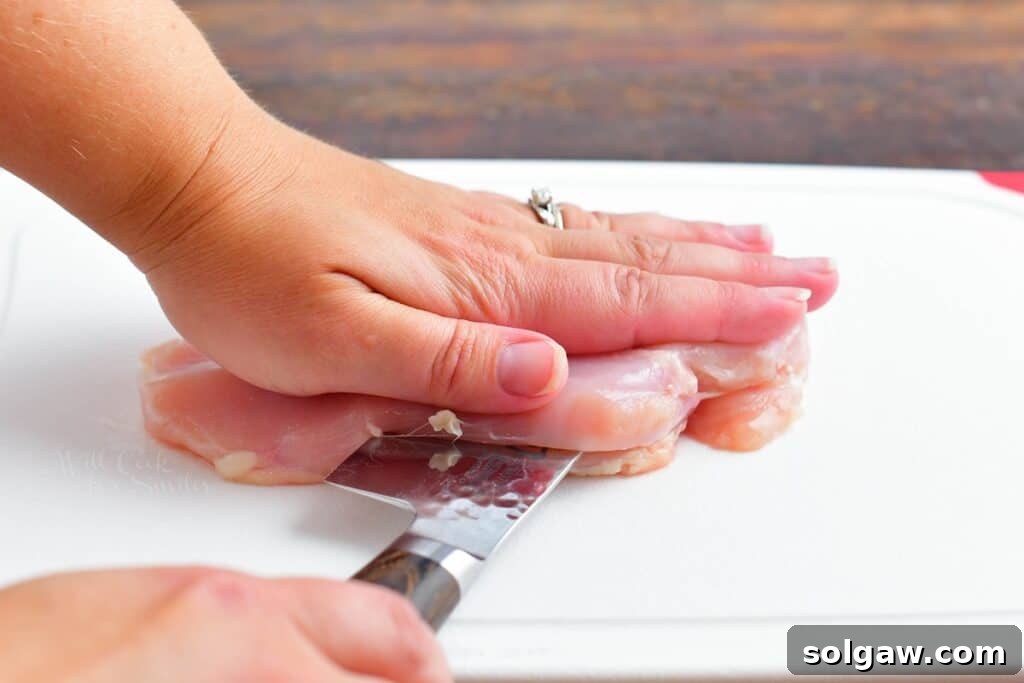 A hand is pressing raw chicken down while a knife is slicing through the middle of the breast.