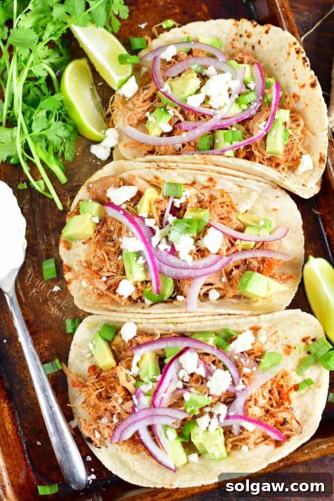 three shredded chicken tacos with toppings on a sheet pan with sour cream and greens next to them
