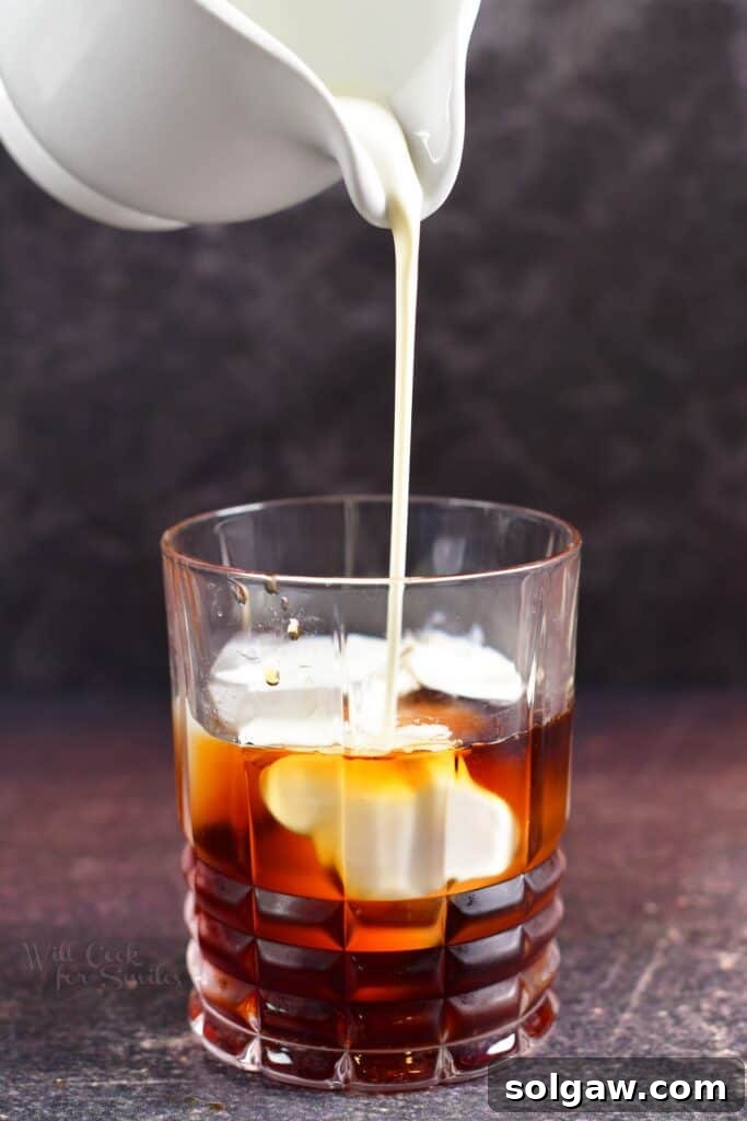 A classic White Russian cocktail being prepared, with heavy whipping cream gently poured over Kahlúa and vodka, creating a beautiful swirling effect.