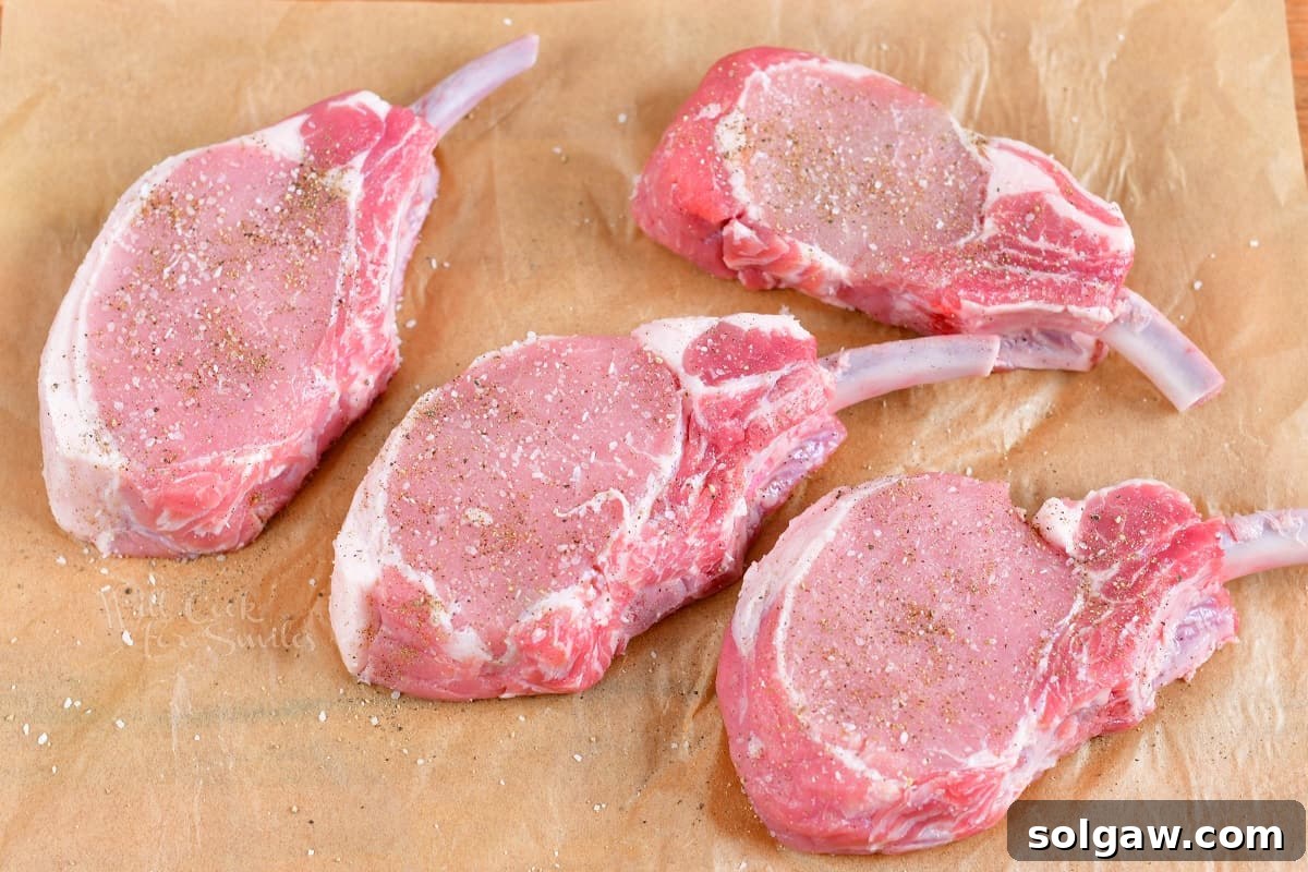 Four thick, raw bone-in pork chops are neatly arranged on a sheet of parchment paper, indicating they are prepared and waiting for the next step of stuffing or seasoning.