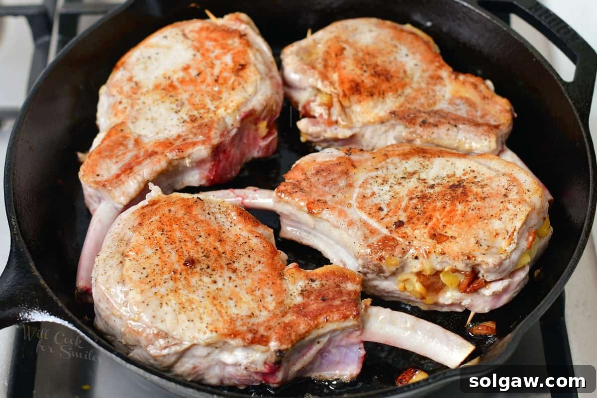Two stuffed pork chops are searing in a hot black pan, developing a delicious golden-brown crust on their surface.