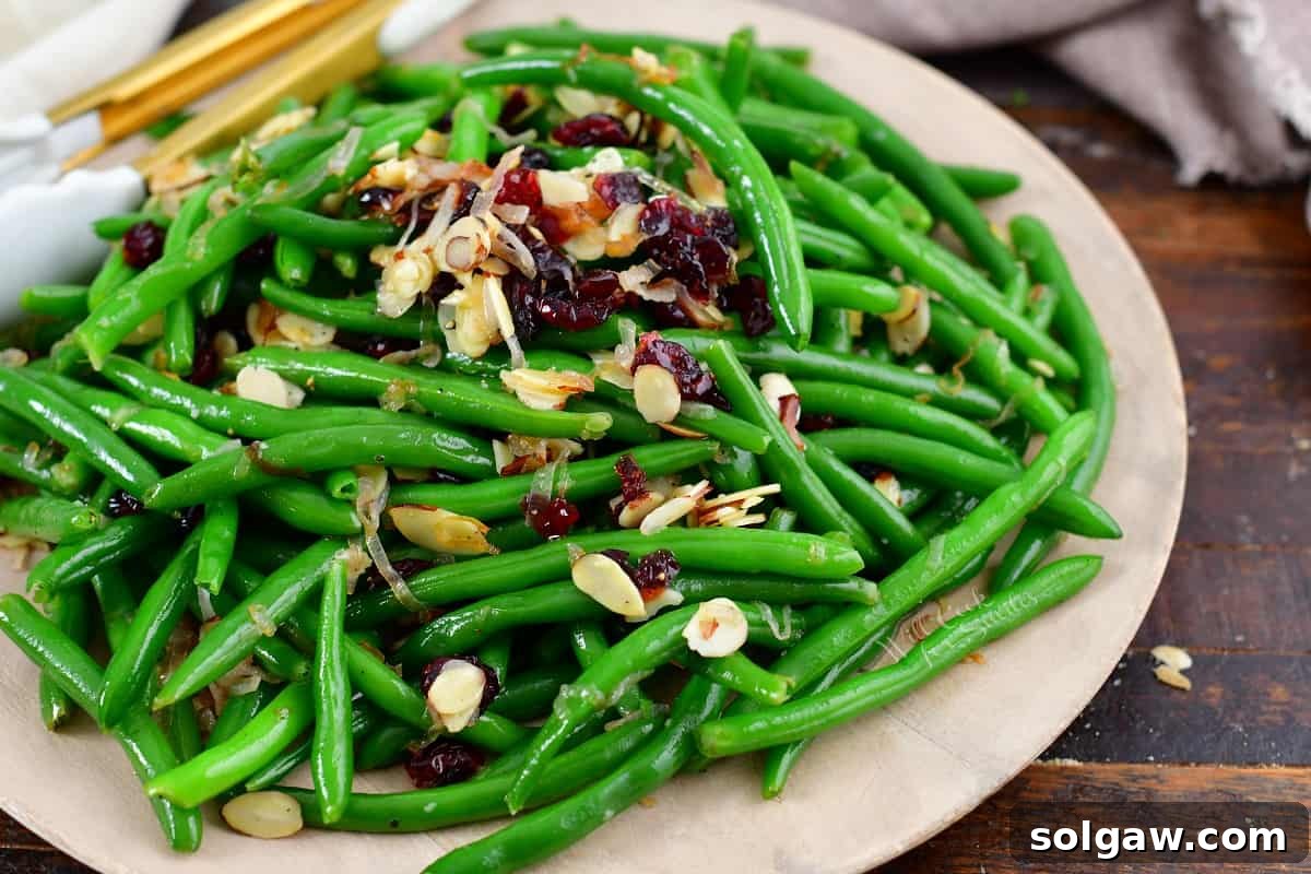 A close-up view of sautéed green beans on a white plate, beautifully garnished with bright red cranberries and slivered almonds, highlighting the dish's vibrant colors and textures.