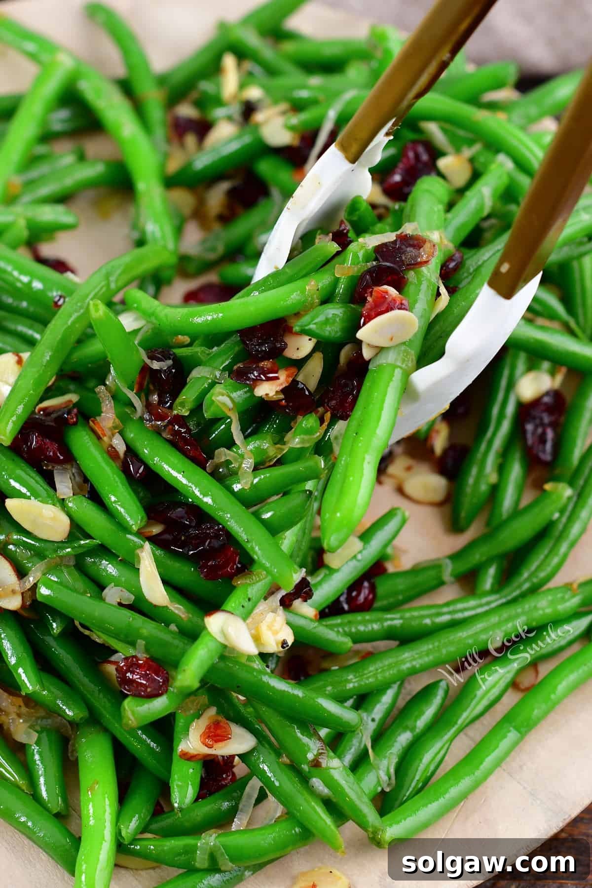 A hand using gold metal tongs to scoop a serving of sautéed green beans from a white serving dish, showing the texture and garnish.