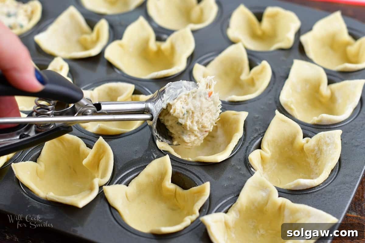 A spoon adding creamy crab filling into the prepared puff pastry cups in a mini muffin pan.