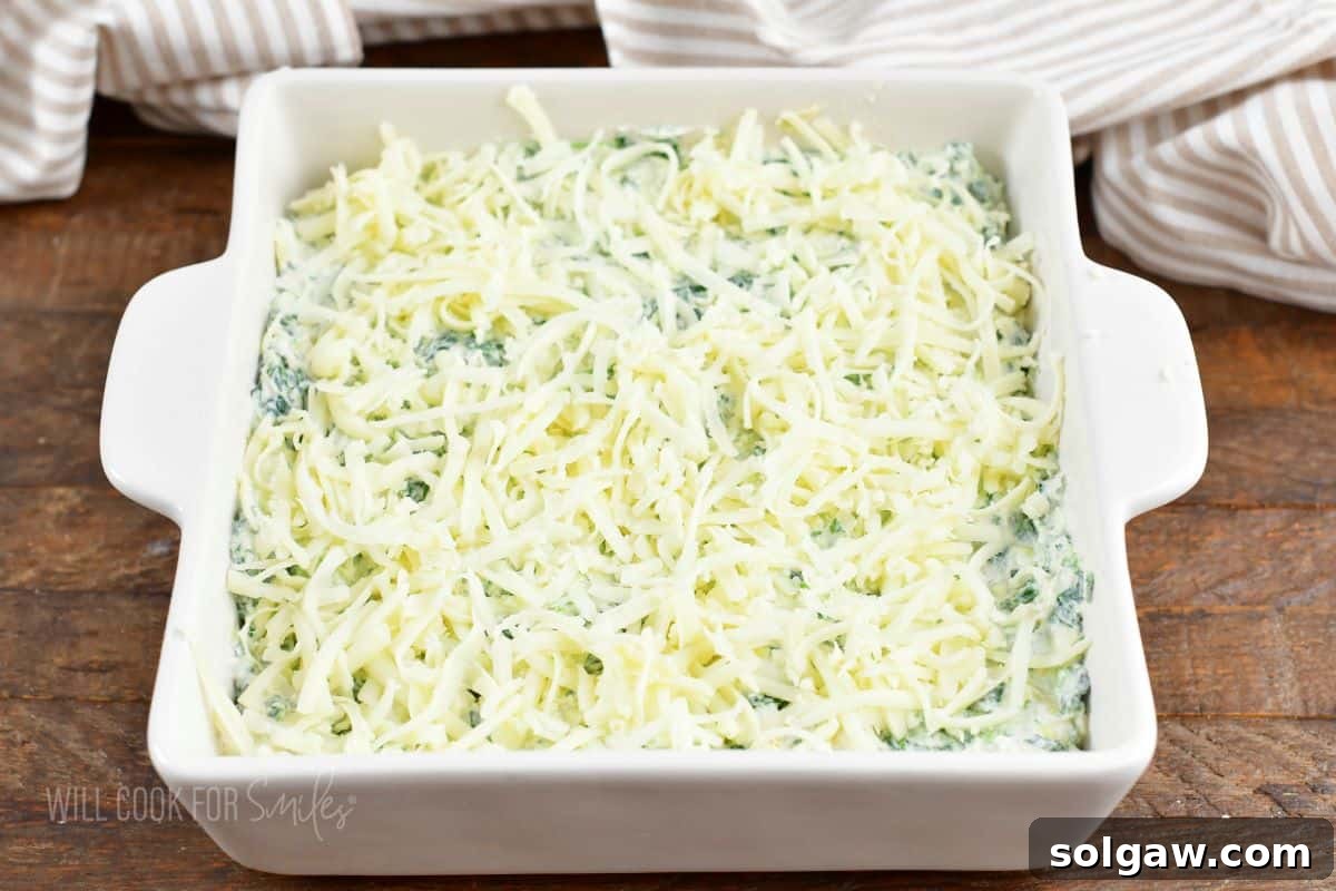 Spinach dip mixture meticulously spread into a pristine white baking dish, generously topped with an abundant layer of shredded cheese, poised for baking.