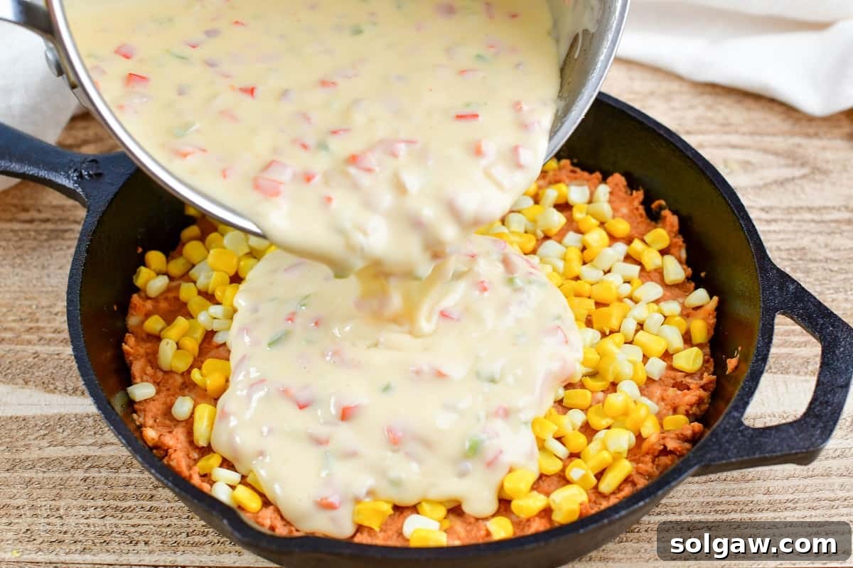 pouring queso mixture over refried bean and corn