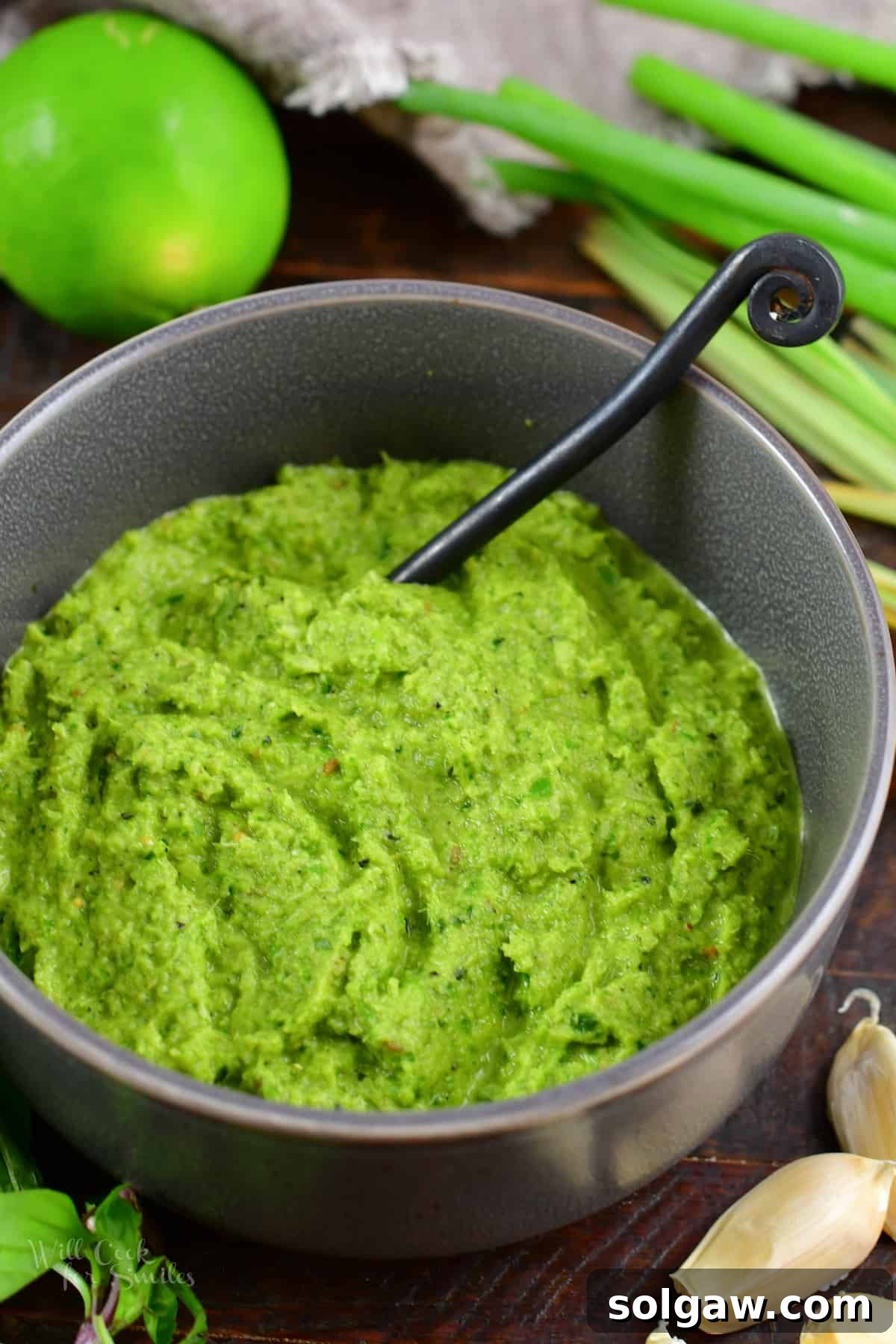 Closeup of vibrant homemade green curry paste in a ceramic bowl with a spoon, surrounded by fresh Thai ingredients.