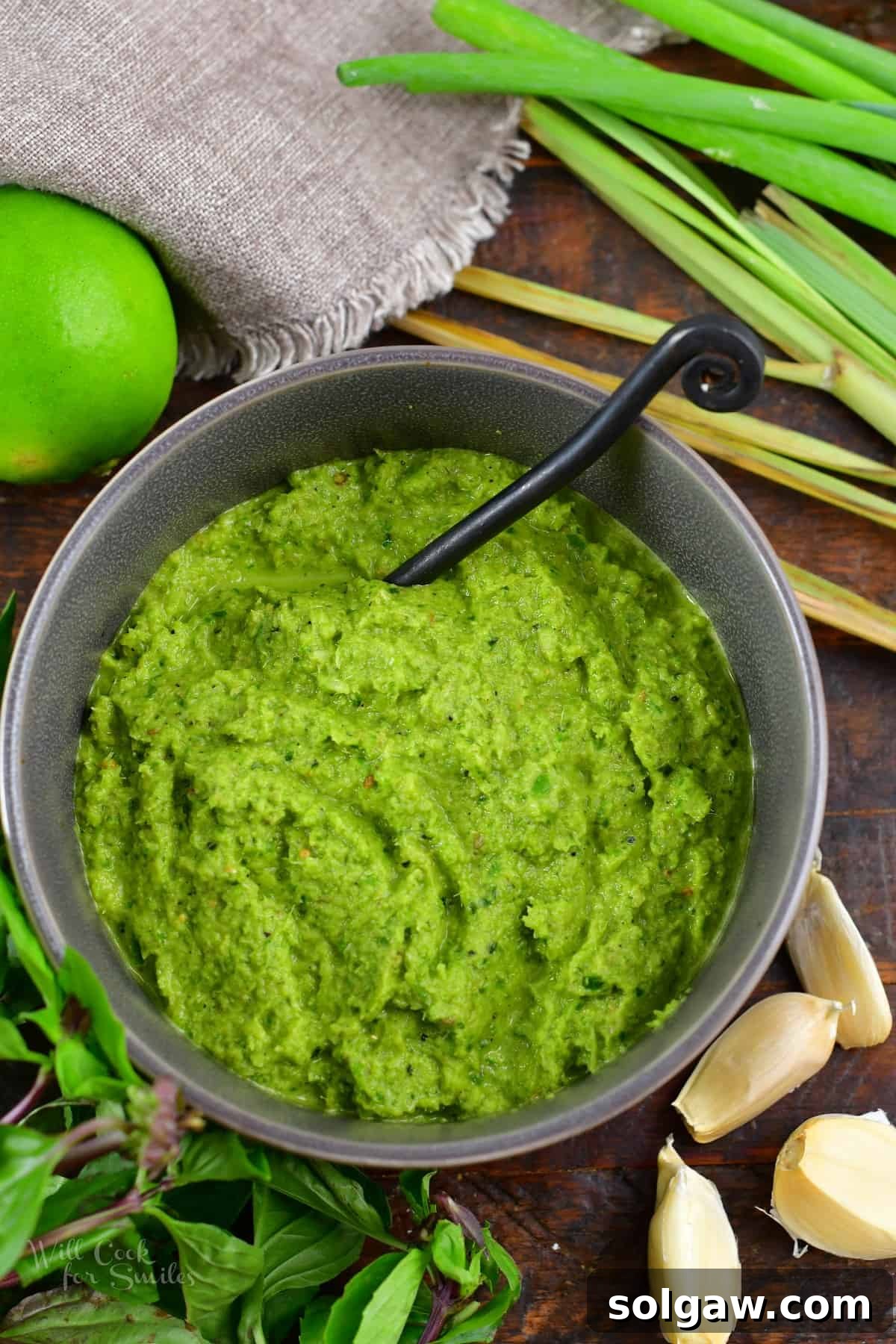 A beautifully arranged top-down shot of green curry paste in a bowl, surrounded by all its fresh, vibrant ingredients on a rustic surface.
