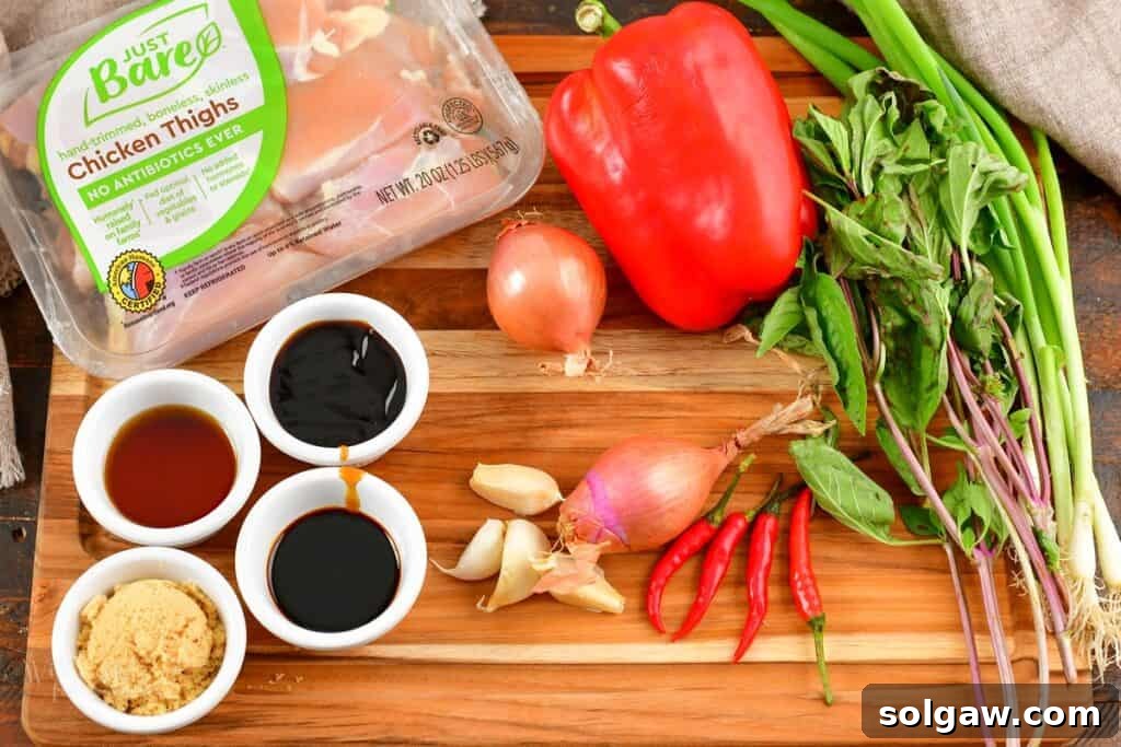 Assortment of fresh ingredients for Thai basil chicken laid out on a wooden board: chicken, garlic, shallots, bell peppers, Thai chilies, and fresh basil leaves.