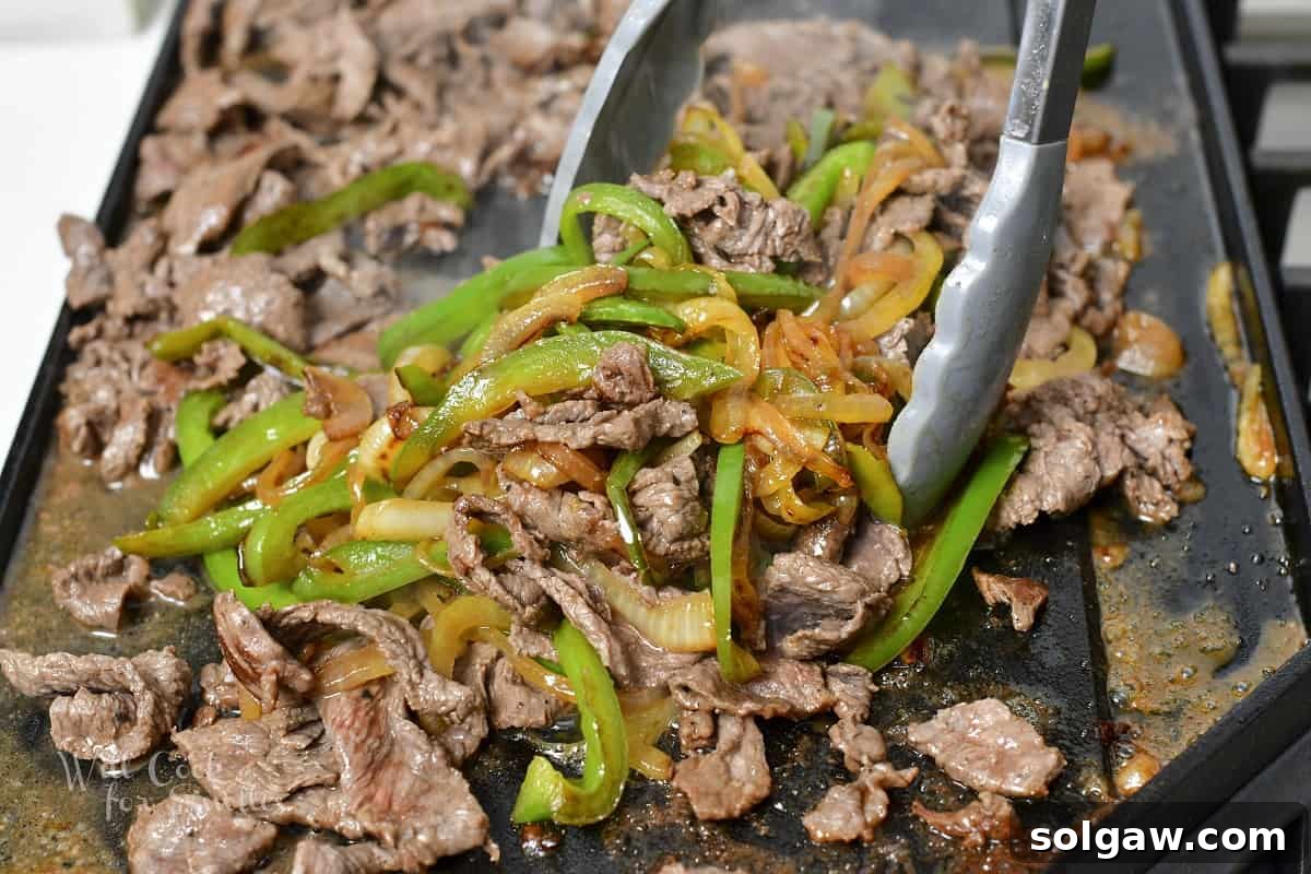 A spatula mixing the cooked onions, peppers, and steaks together on the griddle, ensuring even flavor distribution.