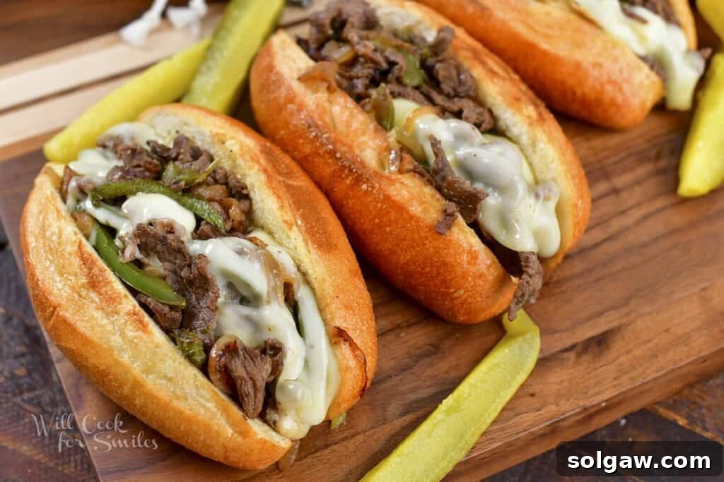 Two perfectly assembled cheesesteak sandwiches, cut in half and served with pickles on a rustic wooden board.