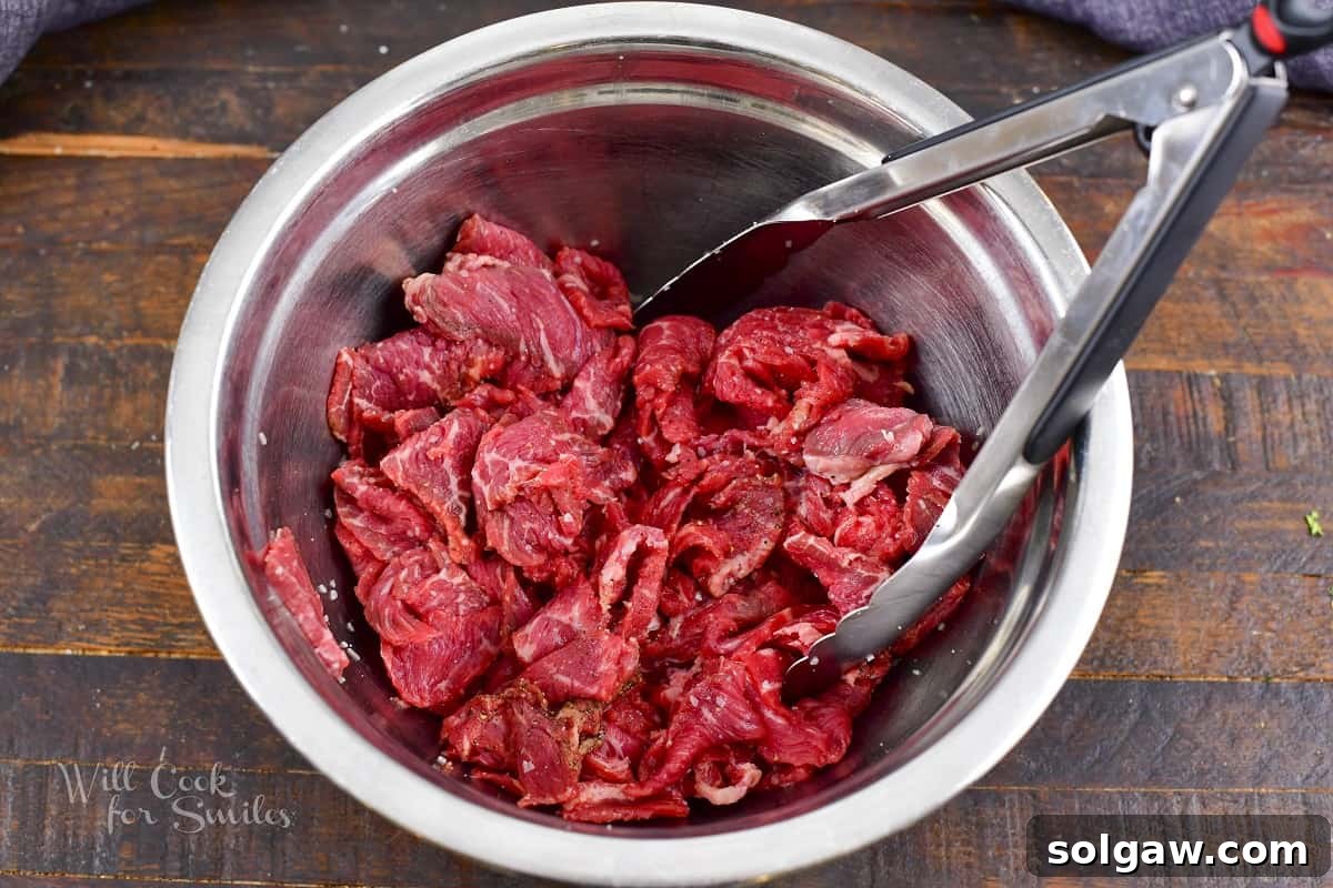 Thinly sliced steak being mixed with seasoning in a bowl, ensuring every piece is coated.