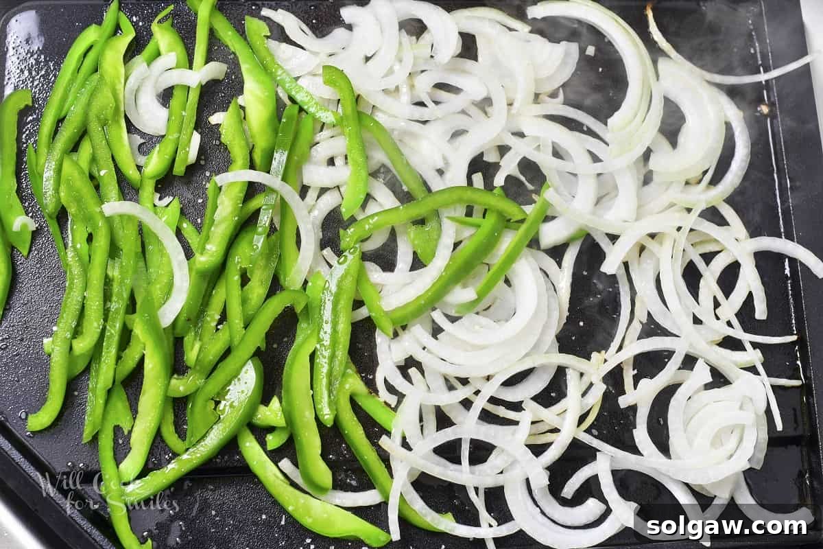 Thinly sliced onions and green bell peppers searing on a hot griddle, starting to caramelize.