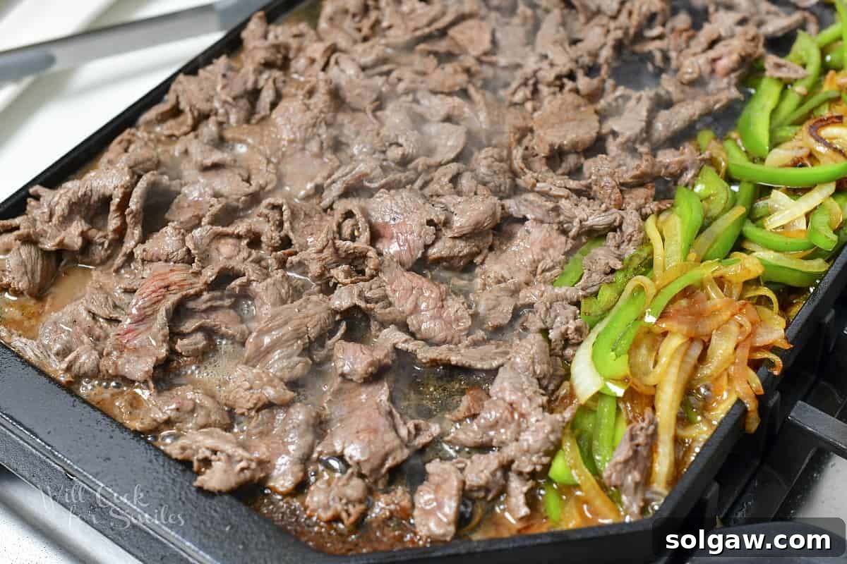 The sizzling steak being expertly seared on the griddle alongside the softening onions and peppers.