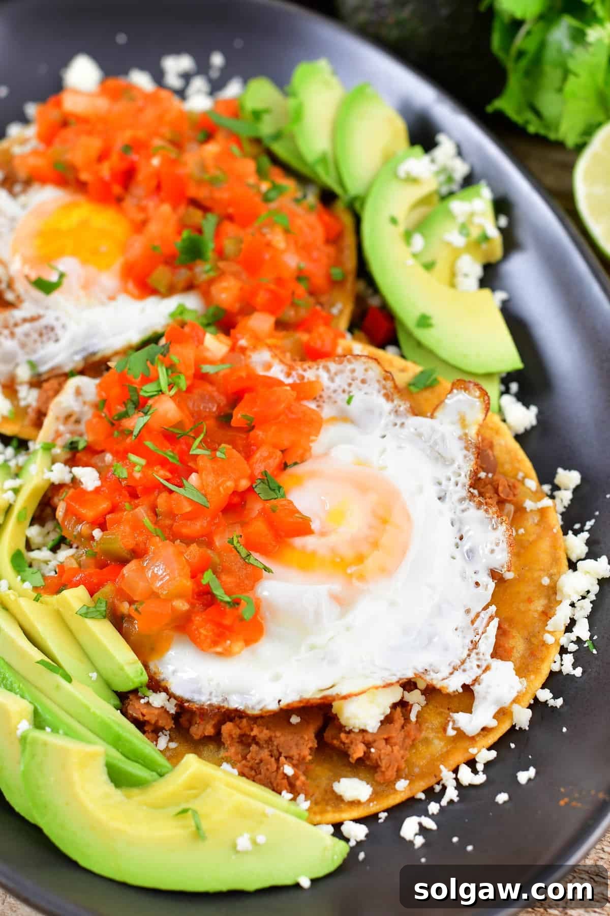Closeup of sunny-side-up eggs over tortillas, refried beans, and avocado, garnished with cilantro and salsa.